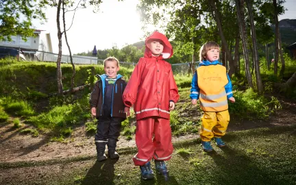Tre barn med regntøy i Fjellvegen barnehage en sommerdag med grønt gress og skog