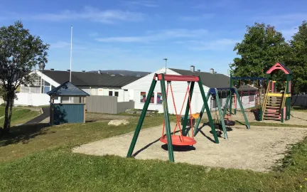 Uteområde i Kroken barnehage med husker, klatretårn, sandkasse, grønt gress og trær en sommerdag med blå himmel 