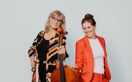 To damer ser i kamera. Den til venstre holder en cello. Foto.