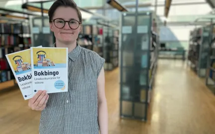 Kvinne med kort brunt hår, briller og grå-og-hvit-stripete skjorte holder to ark og smiler mot kamera. I bakgrunnen er vinduer og bokhyller. Foto.
