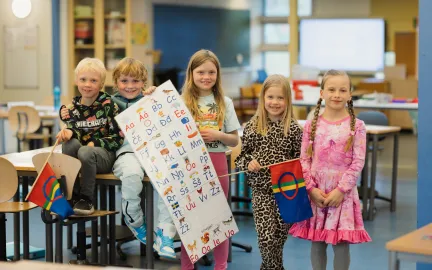Fem barn i barneskolealder står i klasserommet. To av dem har sameflagg i hendene. Midterste barn holder opp en plakat med samiske bokstaver. Alle smiler mot kamera.