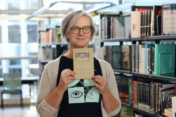 Ellen 150-årsutstilling Kvinne med briller og grå cardigan holder opp en gammel bok og smiler mot kamera. Foto