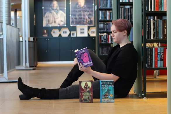 Kvinne sitter på gulvet og leser ei bok med ryggen lent mot ei bokhylle. Hun er kledd i svart og har lange, svarte støvletter på seg. Foran henne står to bøker med fronten mot kamera. Foto.