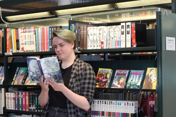 Kvinne i rutete skjorte står lent mot en bokhylle og leser i ei bok. Foto.