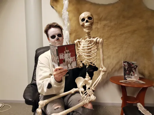 Kvinne med Halloweensminke i ansiktet sitter med et leke-skjelett på fanget. Hun holder opp et filmdeksel. Foto