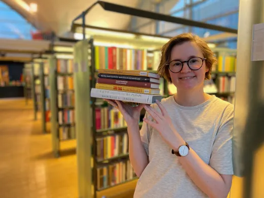 Under radaren uke 46 Kvinne med halvlangt, brunt hår og lys grå t-skjorte holder en bunke bøker og smiler mot kamera. I bakgrunnen står reoler med bøker. Foto