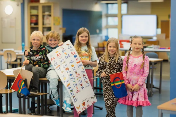 Samiske barn tilhørende Prestvannet skole, fotografert i mai 2025 Fem barn i barneskolealder står i klasserommet. To av dem har sameflagg i hendene. Midterste barn holder opp en plakat med samiske bokstaver. Alle smiler mot kamera.